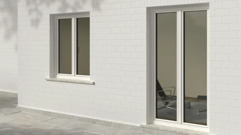Facciata esterna di un edificio con pareti in mattoni bianchi, su cui sono installate due finestre in PVC Finstral: una finestra a due ante e una porta-finestra scorrevole. I serramenti sono di colore chiaro e presentano un design moderno e minimale. All'interno si intravede un ambiente arredato con sedie da ufficio.